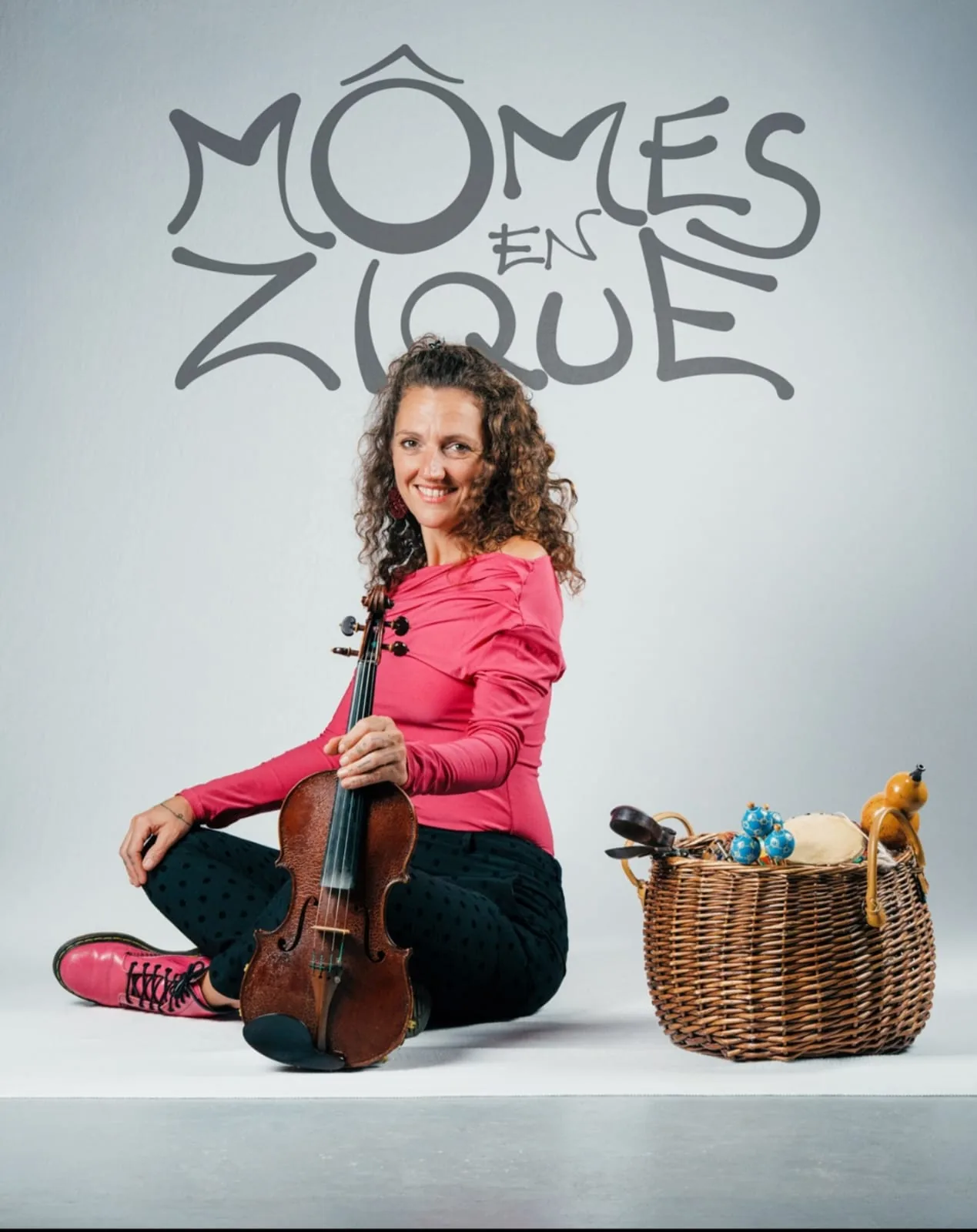 Barbara Lamothe assise au sol avec son violon, à côté d'un panier de petits instruments, devant le logo Mômes en Zique.