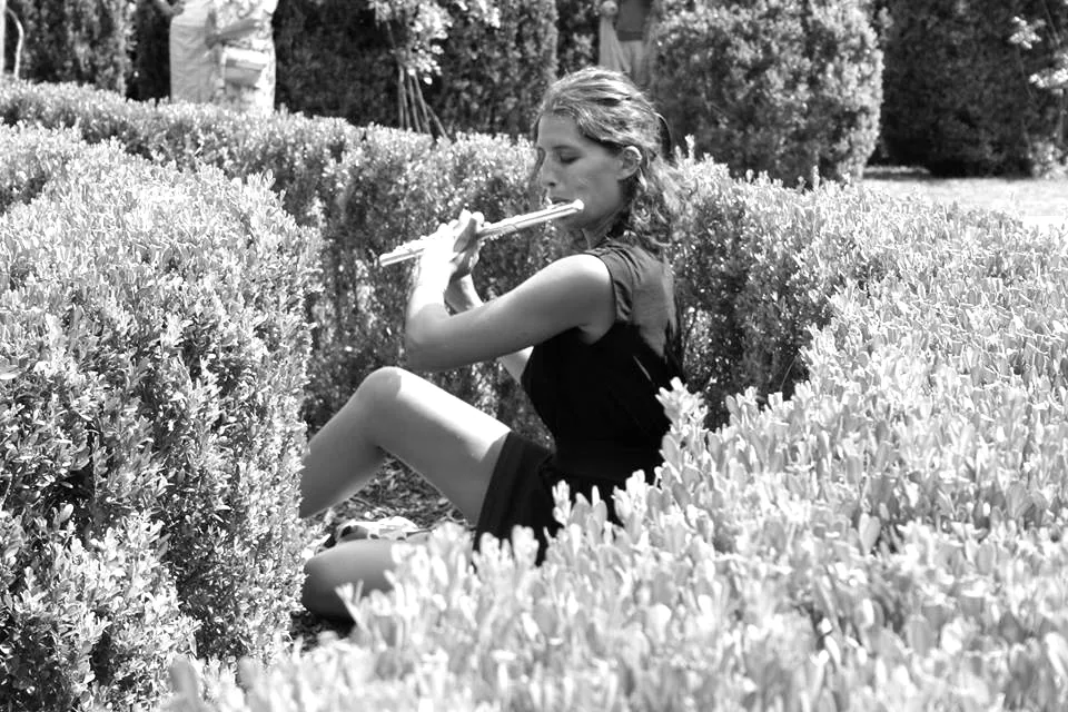 Photo en noir et blanc d'Elisa Lefelle jouant de la flûte traversière, assise au milieu de massifs fleuris.
