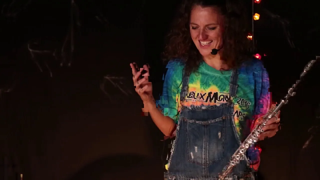 Sur scène, une musicienne en salopette sourit, une flûte traversière à la main.